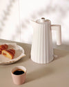 White ribbed electric kettle by Alessi, displayed on a beige counter next to a pink cup of coffee and croissants.