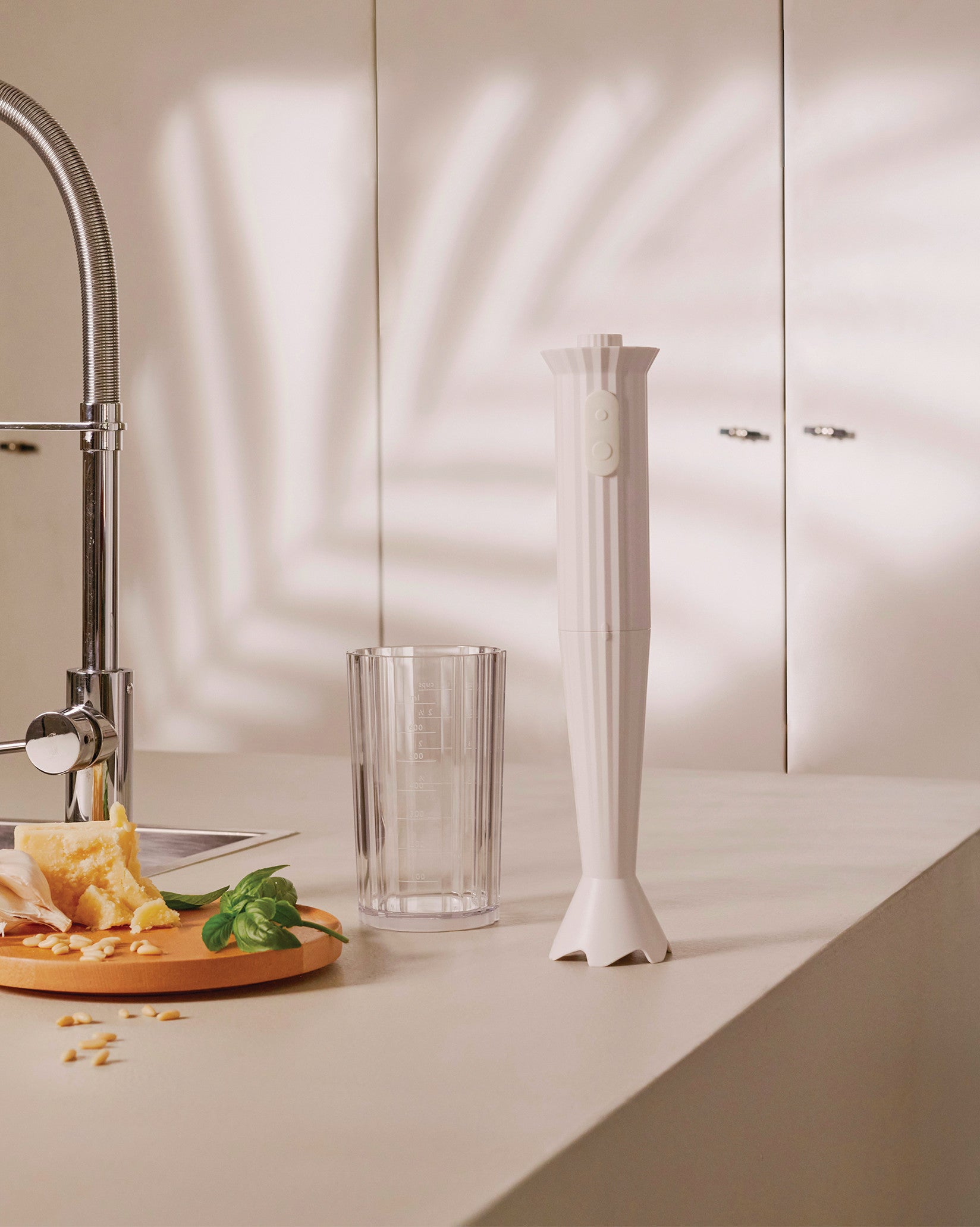 White pleated immersion blender by Alessi standing on a kitchen counter, next to basil, Parmesan, and a measuring cup.