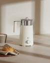 White pleated milk frother by Alessi with a stainless steel pitcher and clear lid.