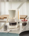 White pleated filter coffee maker by Alessi, brewing dark coffee into a glass carafe, with a digital display showing 7:10.