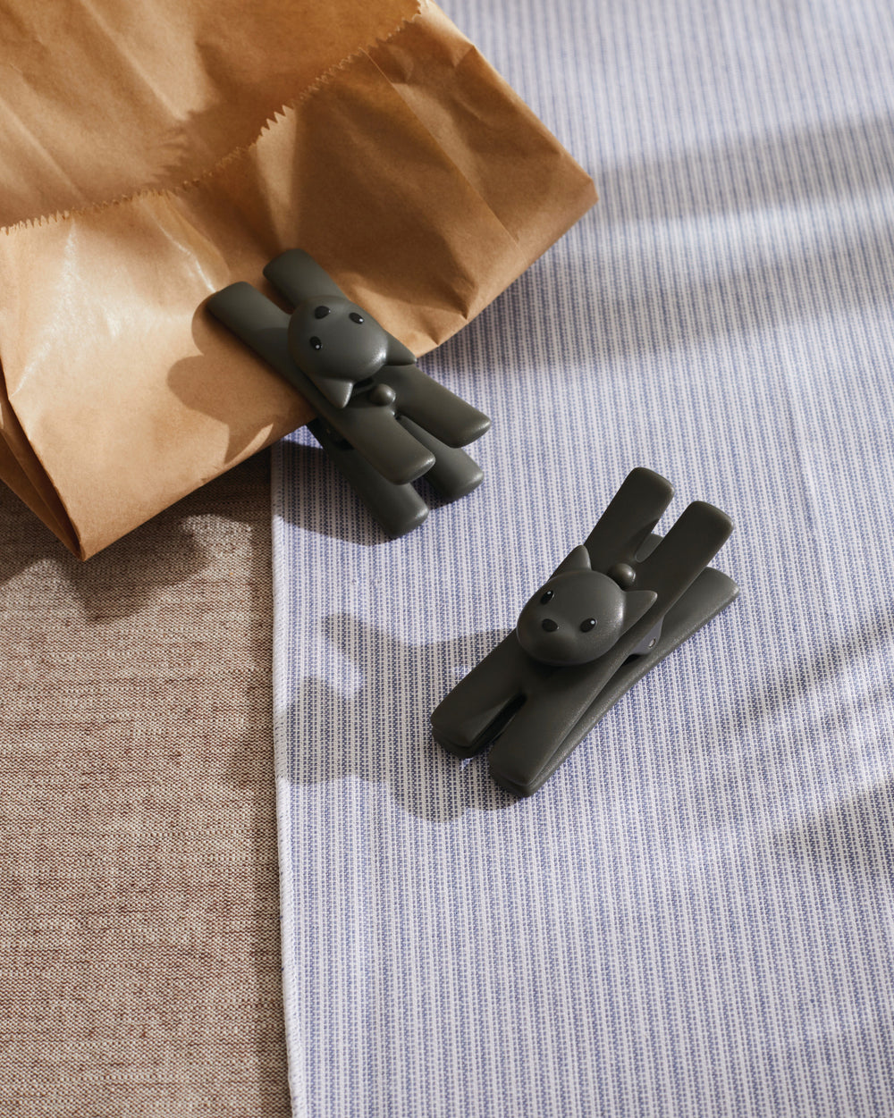 Dark grey cat-shaped bag clips by Alessi, one holding a brown paper bag on blue striped cloth.
