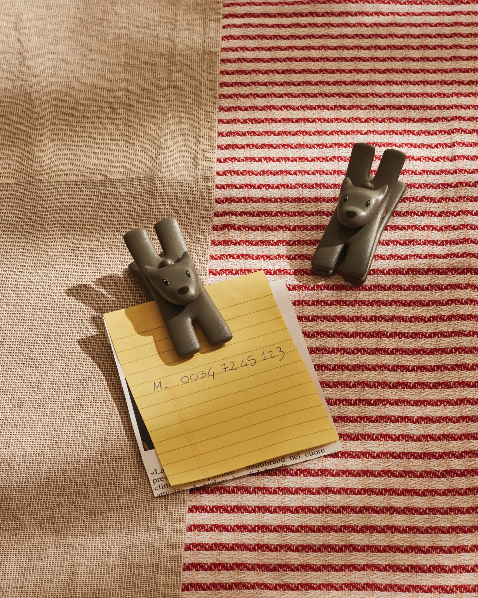 Dark gray dog-shaped Giampo clips by Alessi, one holding a yellow notepad, on a red and beige striped towel.