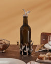 A dark brown glass oil cruet with shiny silver leaf-patterned stand and stopper by Alessi, on a dining table with bread.