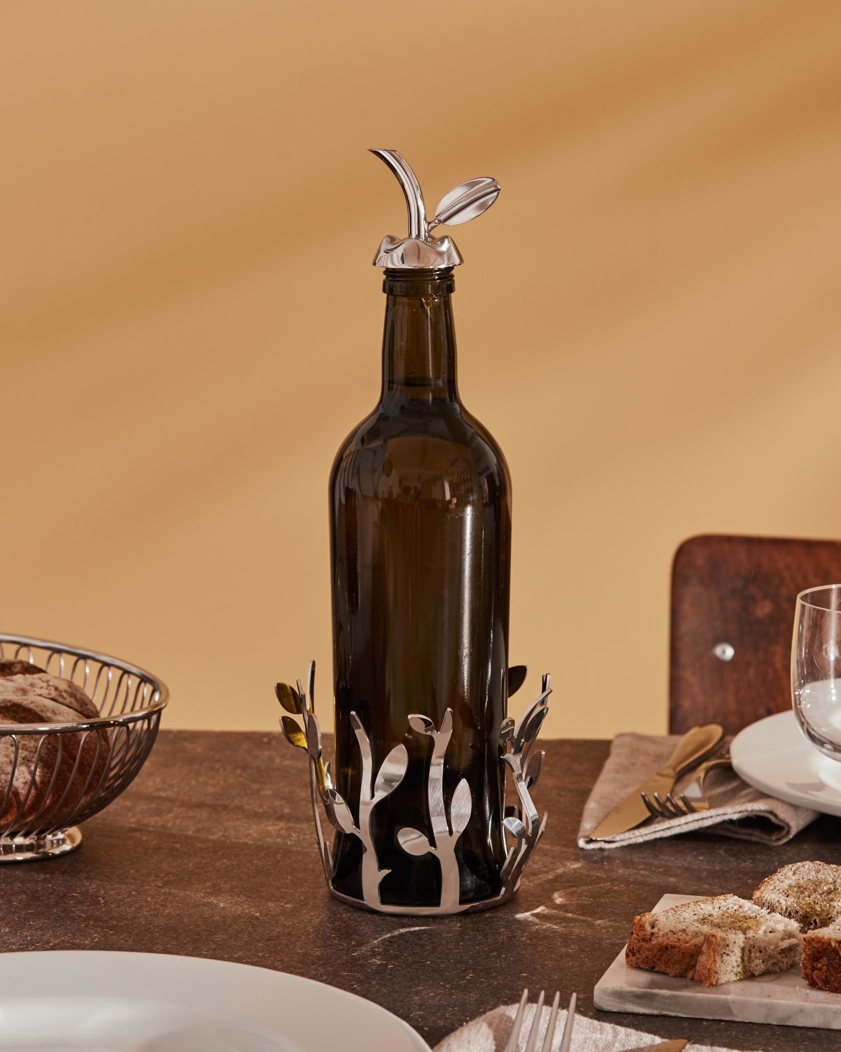 A dark brown glass oil cruet with shiny silver leaf-patterned stand and stopper by Alessi, on a dining table with bread.