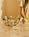 Geometric silver fruit bowl by Alessi filled with green pears on a marble counter, a person's hand holds a pear.