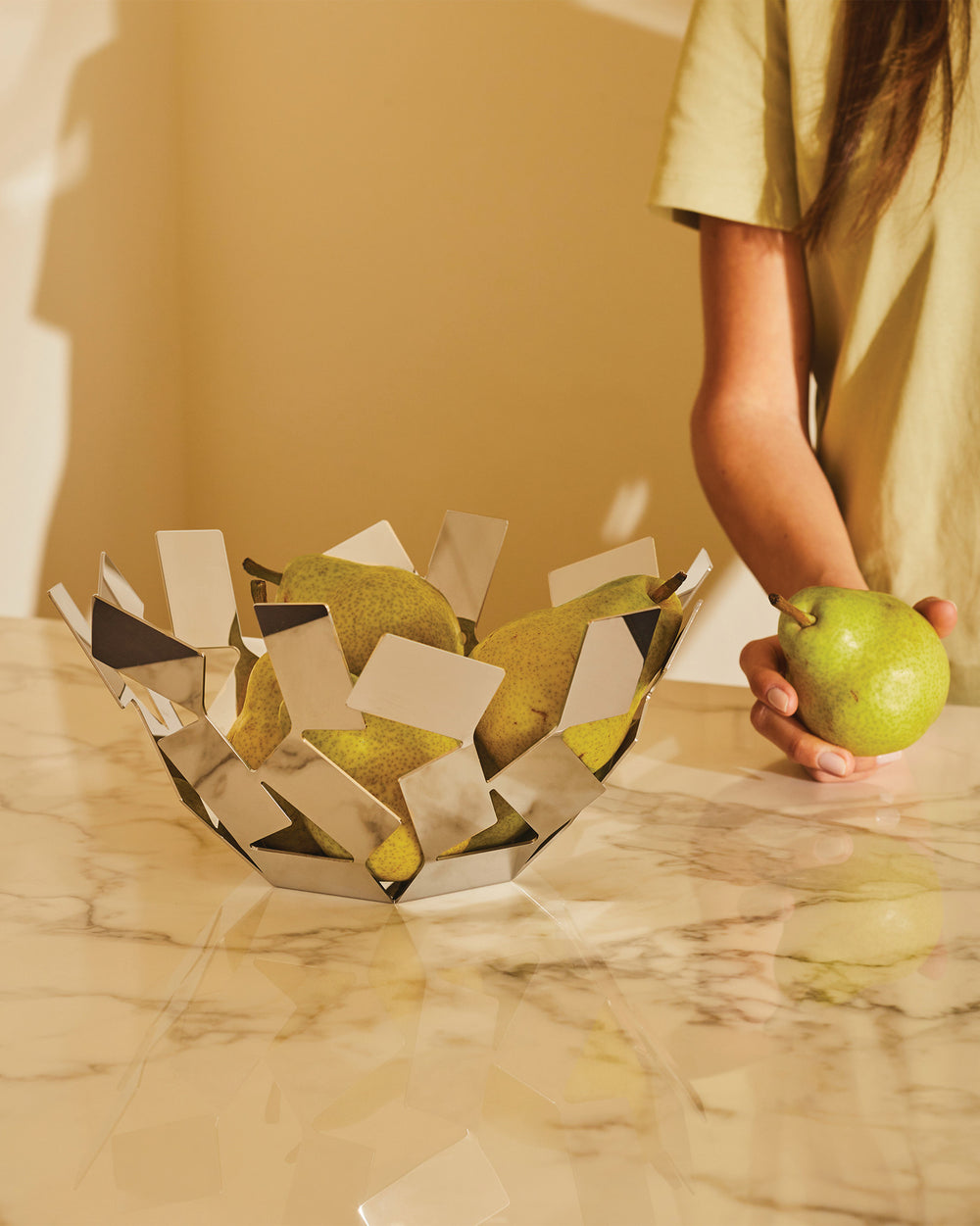 Geometric silver fruit bowl by Alessi filled with green pears on a marble counter, a person's hand holds a pear.
