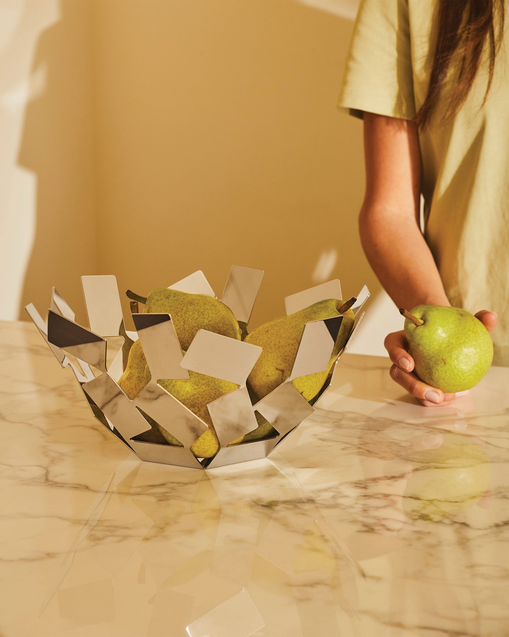 Geometric silver fruit bowl by Alessi filled with green pears on a marble counter, a person's hand holds a pear.