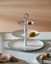 White 3-tier serving stand with scalloped, embossed plates by Alessi, featuring a top handle and colorful macarons.