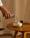 Home fragrance diffuser refill with clear liquid and blue botanical pattern by Alessi being poured into a white ceramic diffuser on a wooden table.