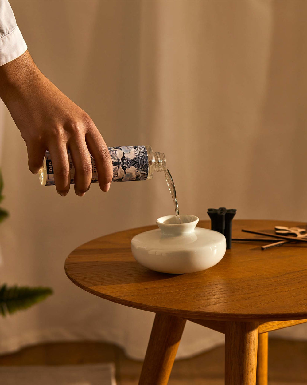 Home fragrance diffuser refill with clear liquid and blue botanical pattern by Alessi being poured into a white ceramic diffuser on a wooden table.