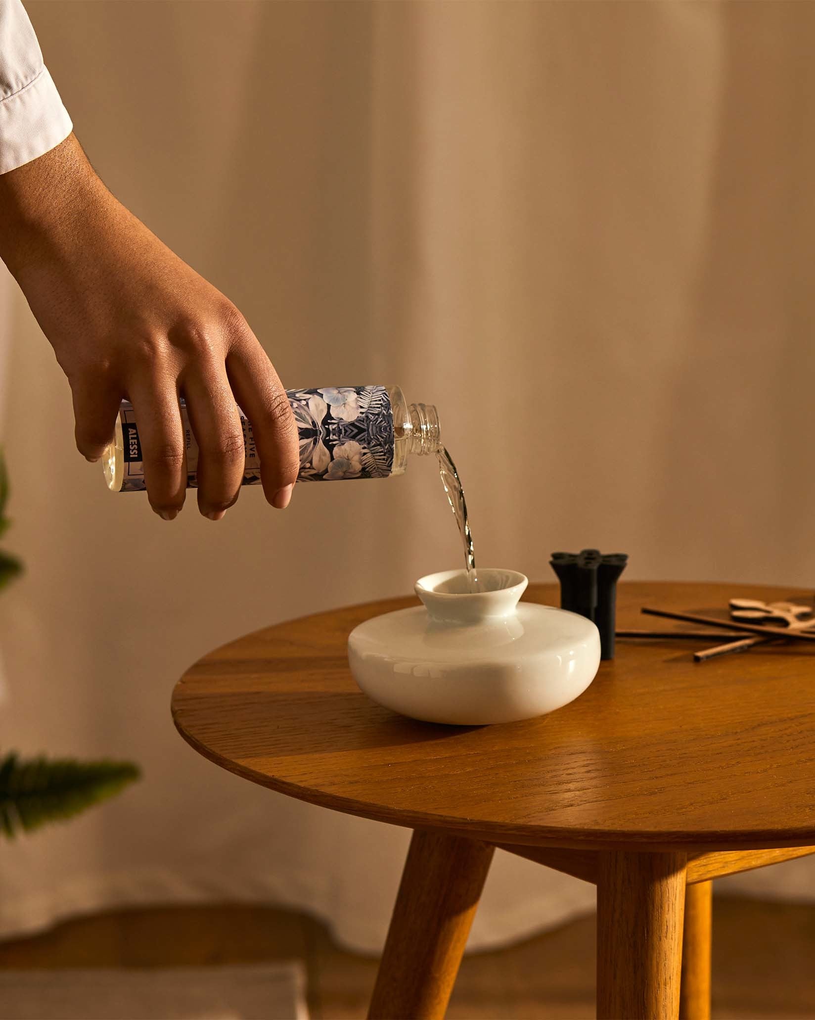 Home fragrance diffuser refill with clear liquid and blue botanical pattern by Alessi being poured into a white ceramic diffuser on a wooden table.