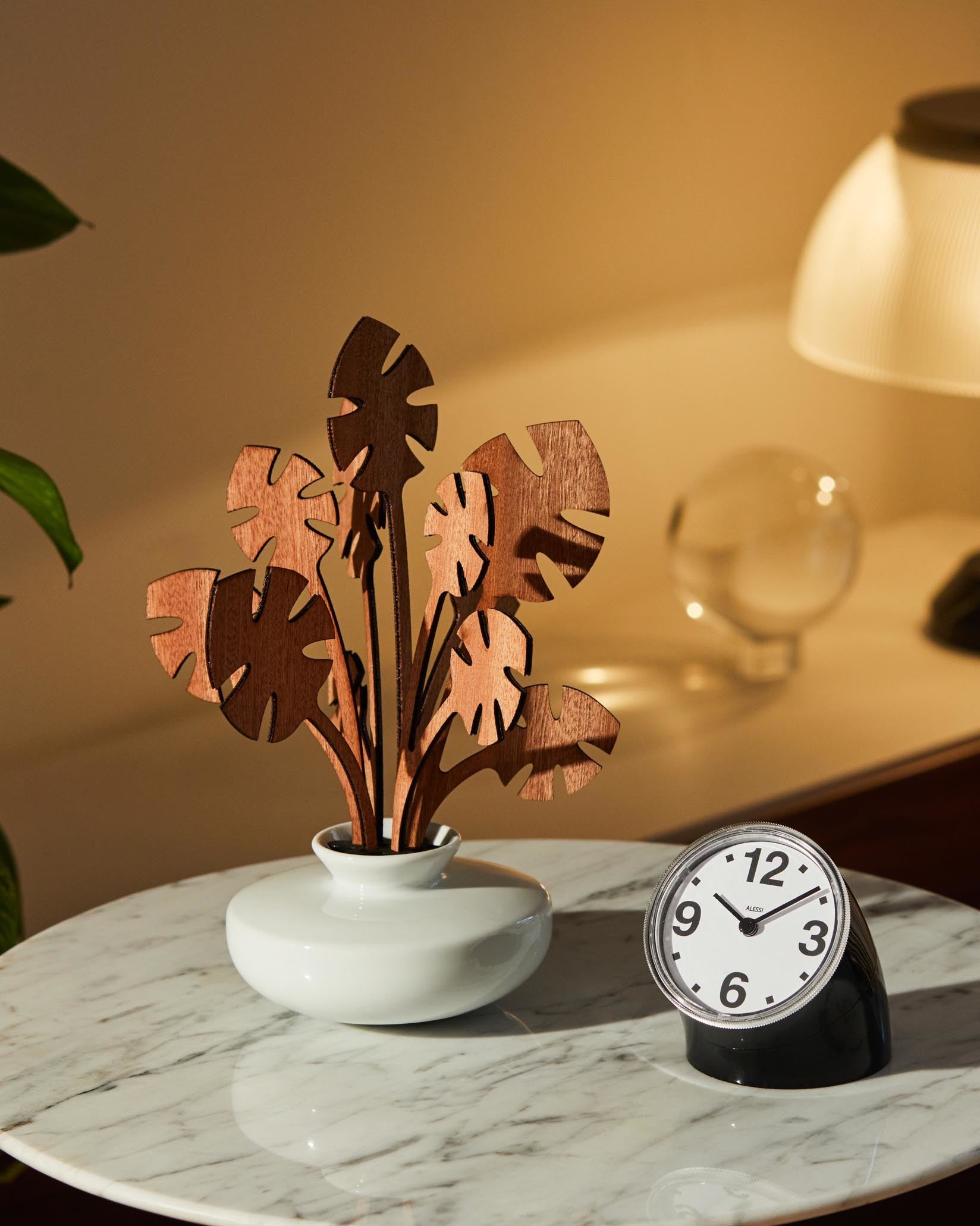 Brown wooden decorative plant by Alessi with abstract leaf designs, in a white vase, next to a black alarm clock on a marble table.