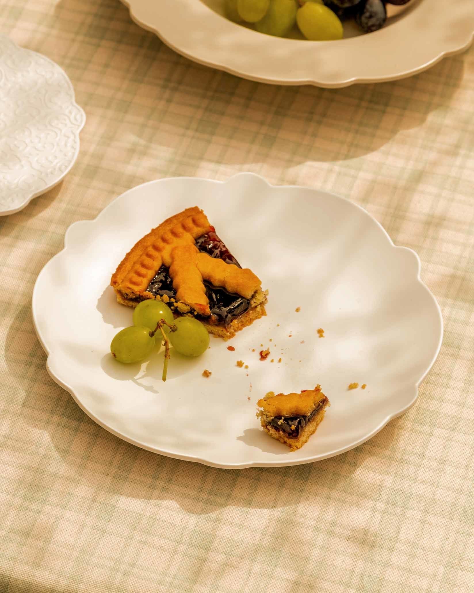 White scalloped plate by Alessi, holding a fruit tart and green grapes on a subtle plaid tablecloth.