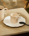 Espresso cup and patterned saucer in light beige ceramic by Alessi, with an ornate black spoon.
