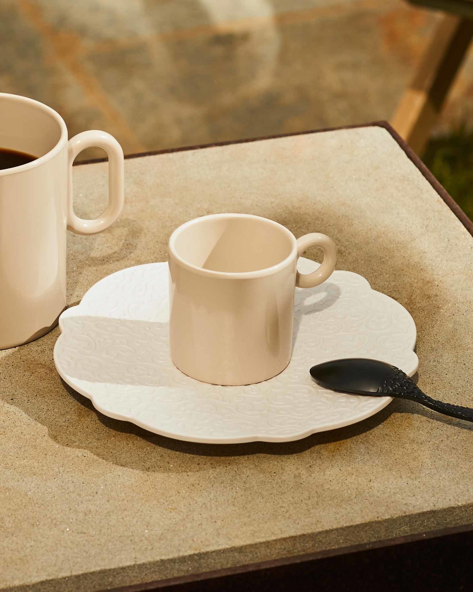 Espresso cup and patterned saucer in light beige ceramic by Alessi, with an ornate black spoon.