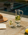 Clear glass carafe by Alessi, filled with water, lemon, cucumber, and mint, with a matching glass.