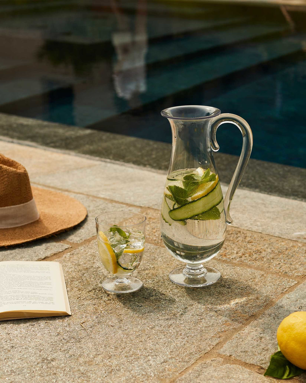 Clear glass carafe by Alessi, filled with water, lemon, cucumber, and mint, with a matching glass.