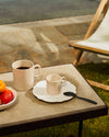 White scalloped plate with relief pattern by Alessi and light beige mugs on a textured outdoor table.