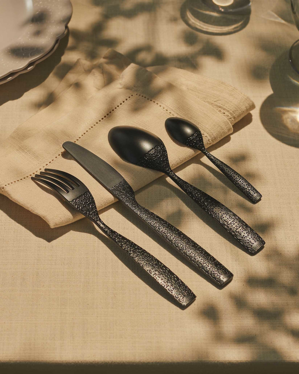Black cutlery set by Alessi featuring ornate textured handles, resting on a beige linen napkin in dappled sunlight.