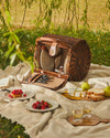 Woven brown picnic basket by Alessi open on a beige blanket with plates, cutlery, and picnic food outdoors.