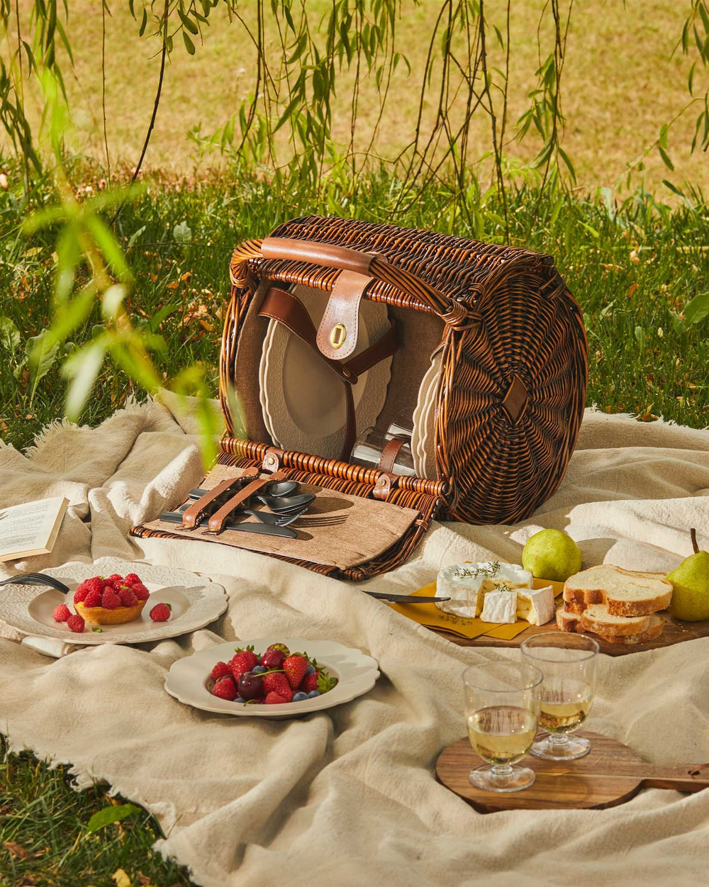 Woven brown picnic basket by Alessi open on a beige blanket with plates, cutlery, and picnic food outdoors.