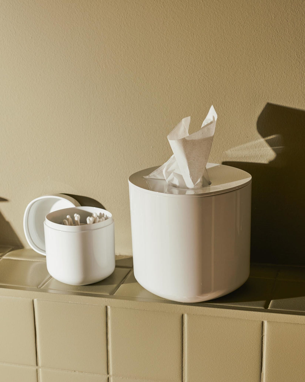 White round tissue dispenser by Alessi, displaying tissues, next to a matching cotton swab holder on green tiles.