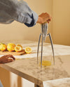 Metallic, three-legged citrus squeezer by Alessi extracting lemon juice into a glass on a marble countertop.