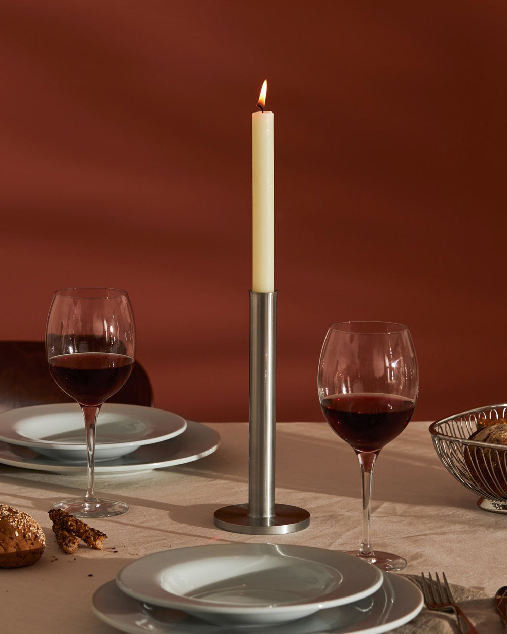 Tall, slender brushed stainless steel candle holder by Alessi, holding a cream taper candle on a set dining table.
