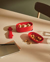 Red food container by Alessi, holding sandwiches, with a separate dish of cookies and notebooks on a beige table.