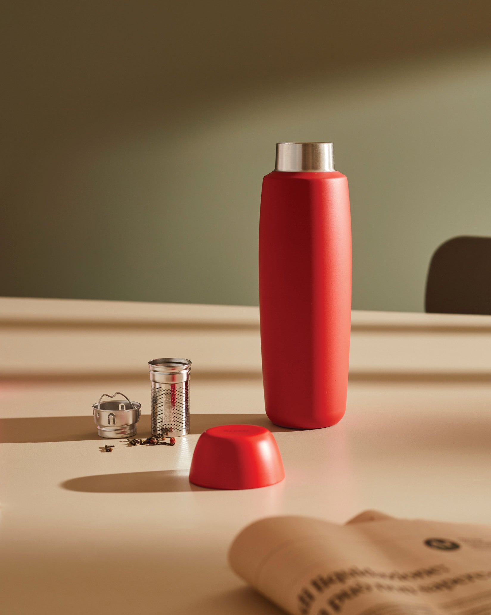 Red thermos bottle by Alessi, standing upright on a beige table next to its red lid, a metal tea infuser, and loose tea leaves.
