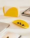 Yellow semi-elliptical desk tray by Alessi, holding paper clips and binder clips on a white surface.
