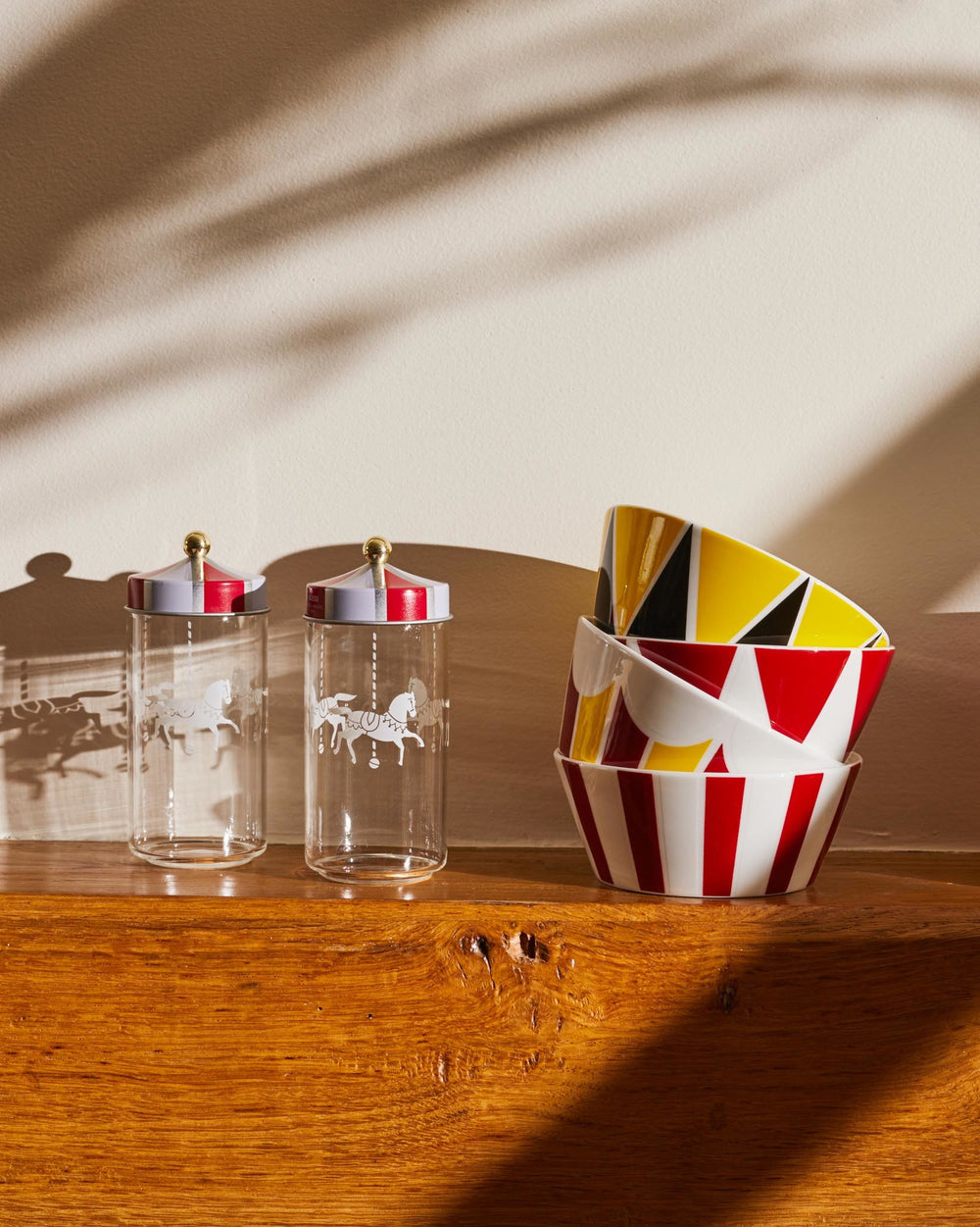 Clear glass jars with conical red and white striped lids by Alessi, featuring white carousel horse designs, displayed with colorful patterned bowls.