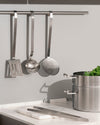 Stainless steel flatware set by Alessi, featuring a spatula, ladle, skimmer, and tongs in a modern kitchen.