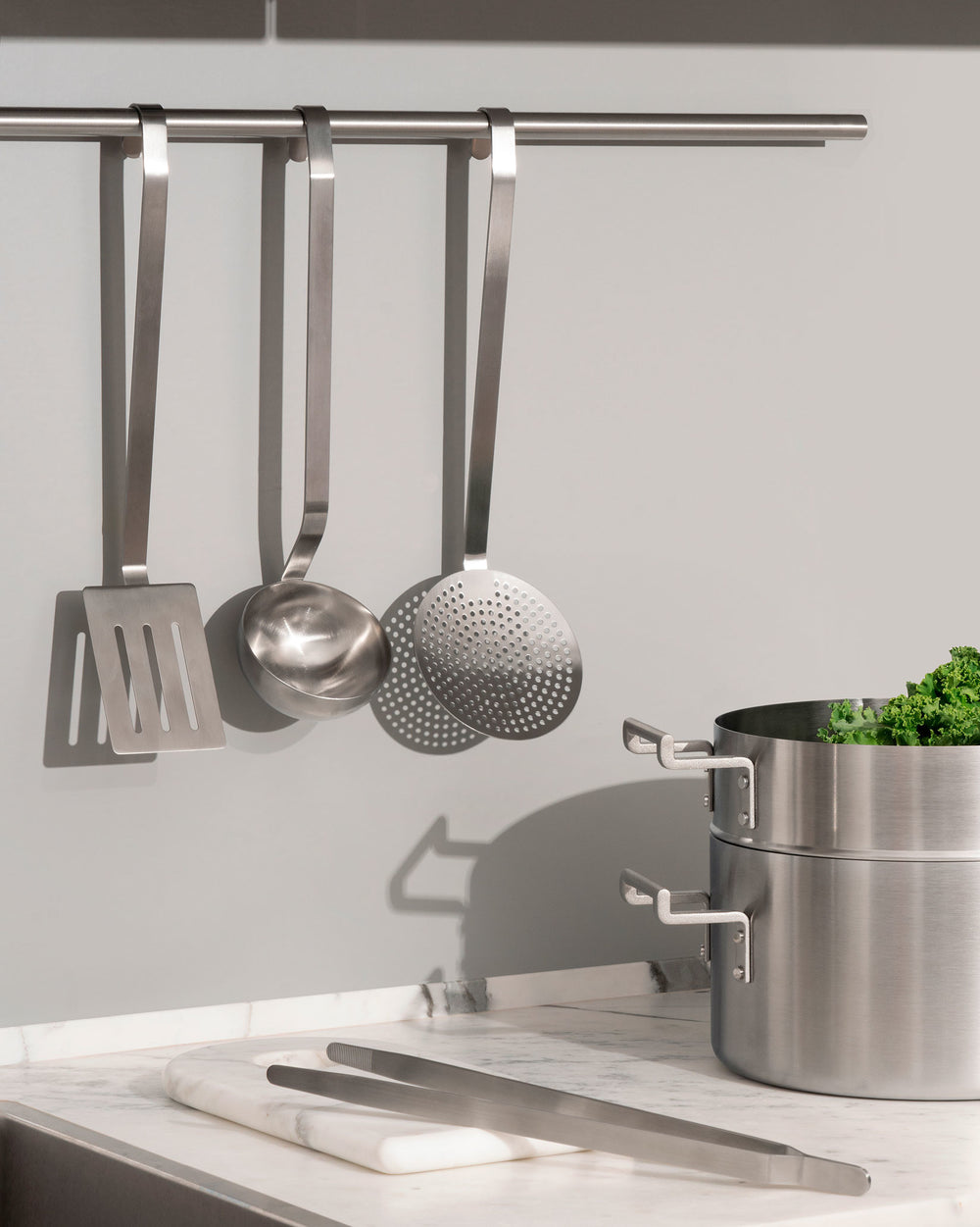 Stainless steel flatware set by Alessi, featuring a spatula, ladle, skimmer, and tongs in a modern kitchen.