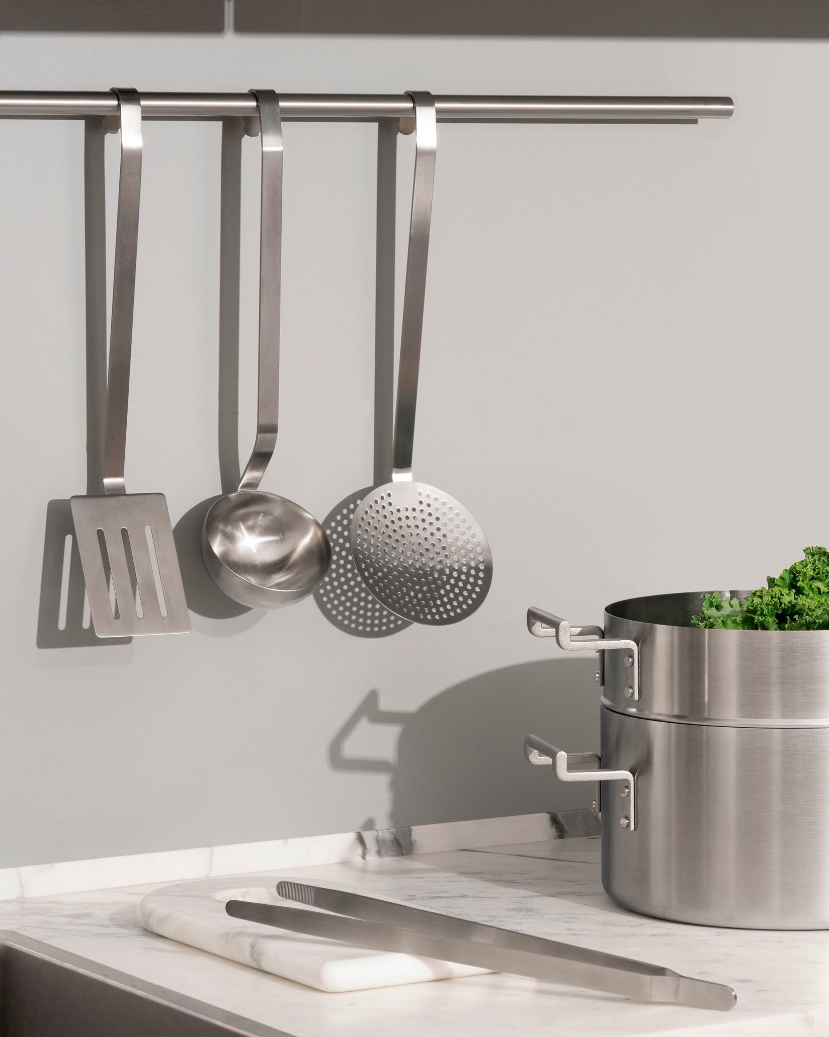 Stainless steel flatware set by Alessi, featuring a spatula, ladle, skimmer, and tongs in a modern kitchen.