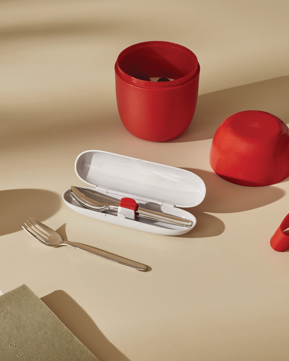 Red portable food carrier and silver cutlery set by Alessi, with a white cutlery case and a notebook on a light tan surface.