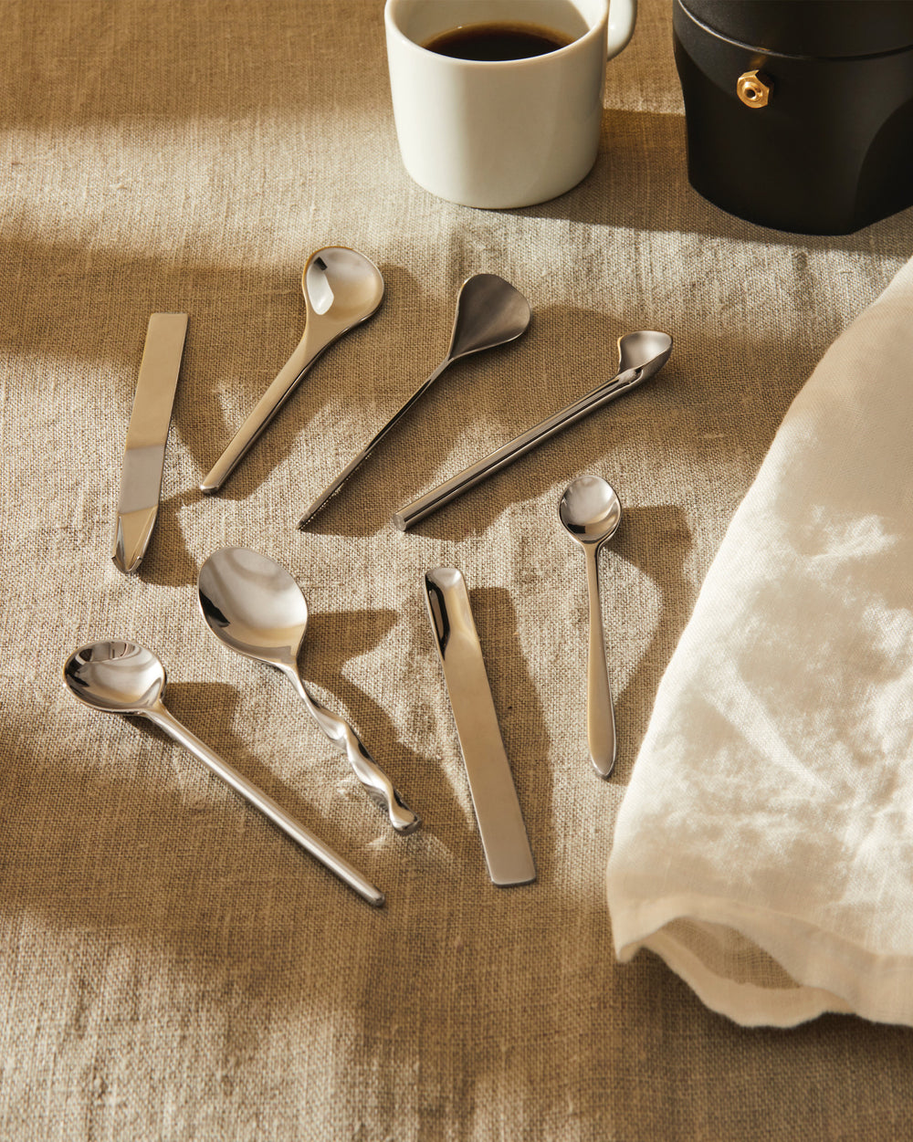 Silver-colored coffee/tea spoons by Alessi, a set of six unique designs on a textured beige tablecloth.