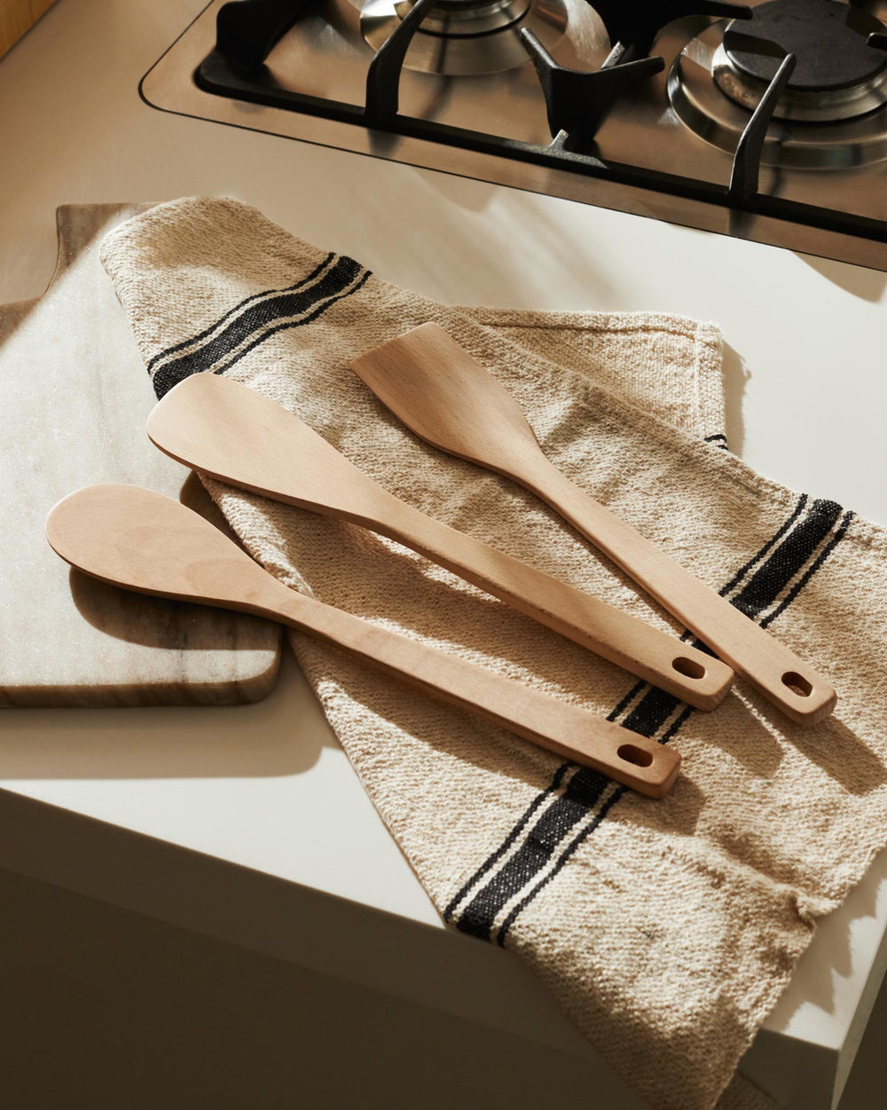 Three light wood kitchen spatulas and spoons by Alessi, with holes in handles, laid on a striped towel on a counter.