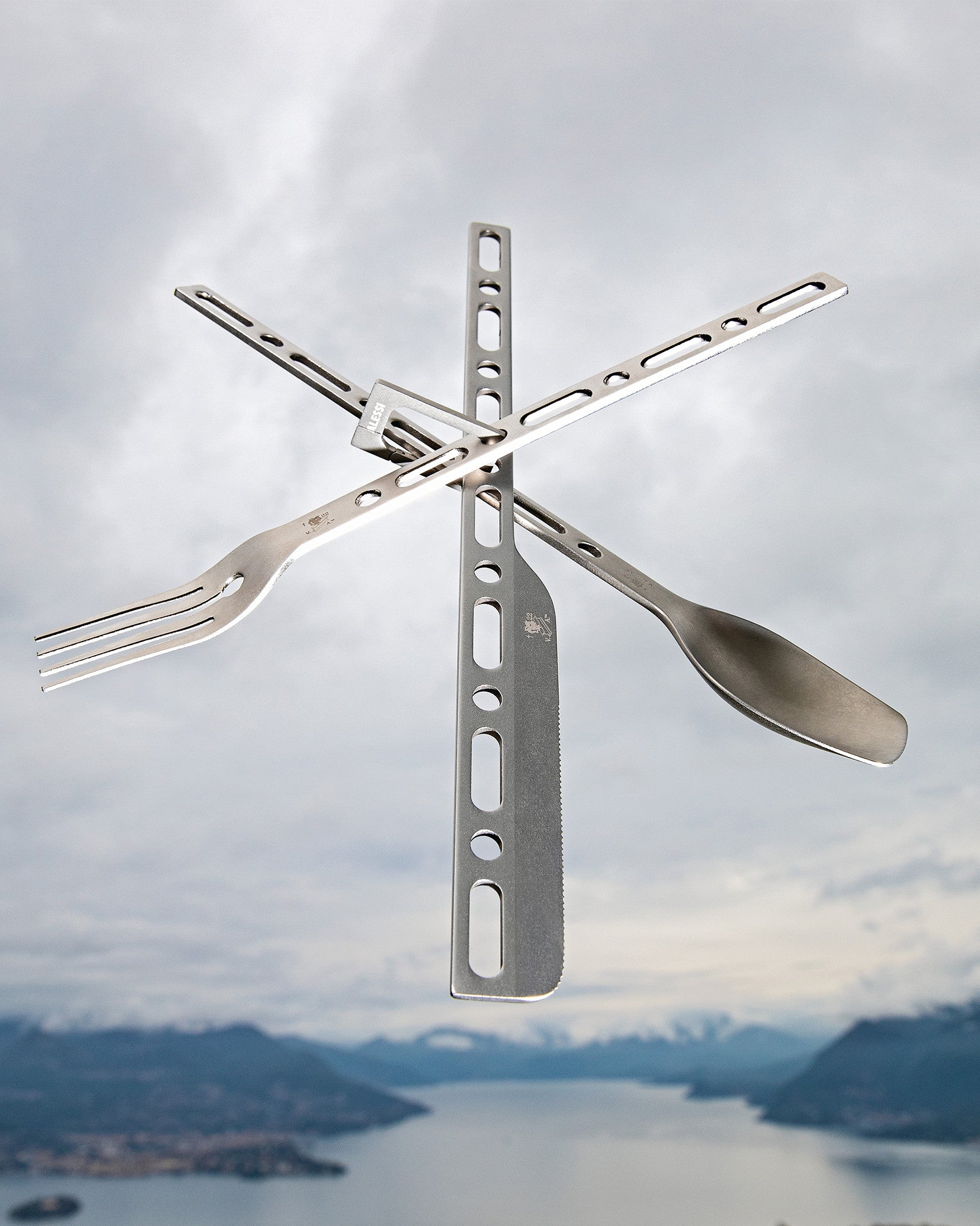 A grey metal modular travel cutlery set by Alessi, with a fork, serrated knife, and spoon arranged in a cross, against a blurred mountain lake.