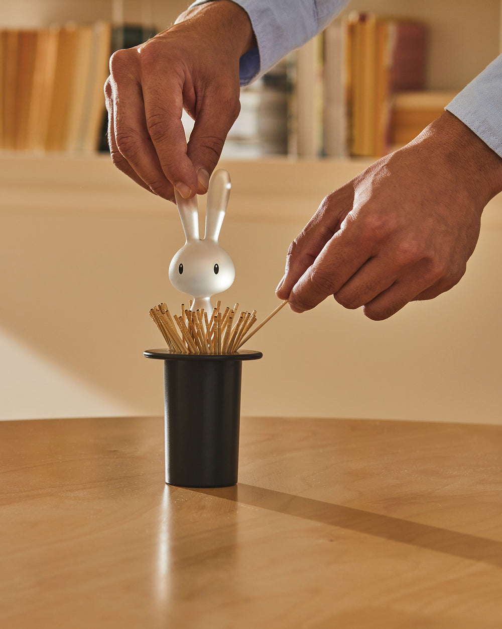 White bunny toothpick holder by Alessi, shaped like a rabbit emerging from a black top hat filled with toothpicks, held by hands.