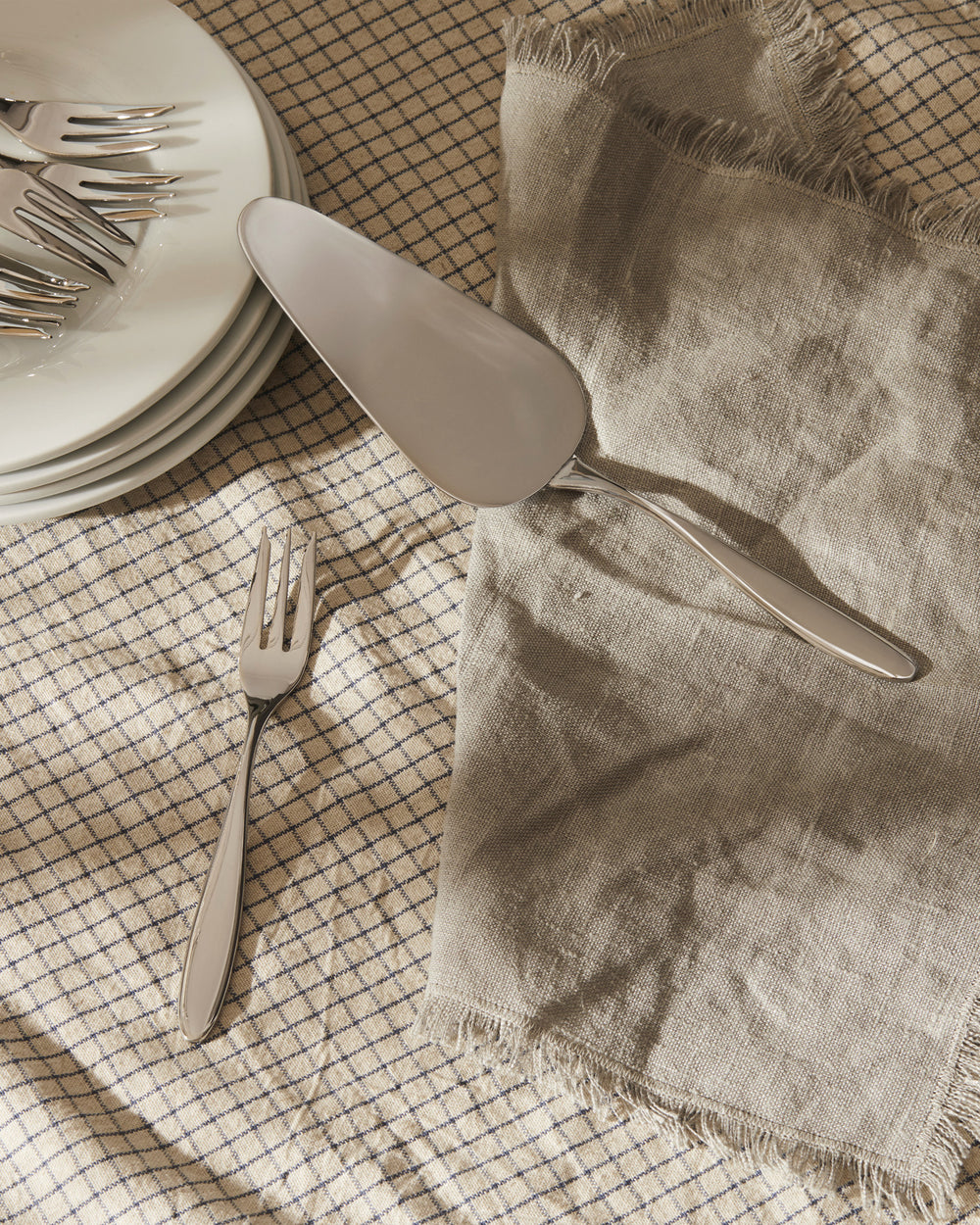 Sleek silver cake server by Alessi and a matching fork, on a checkered tablecloth with a linen napkin and stacked white plates.
