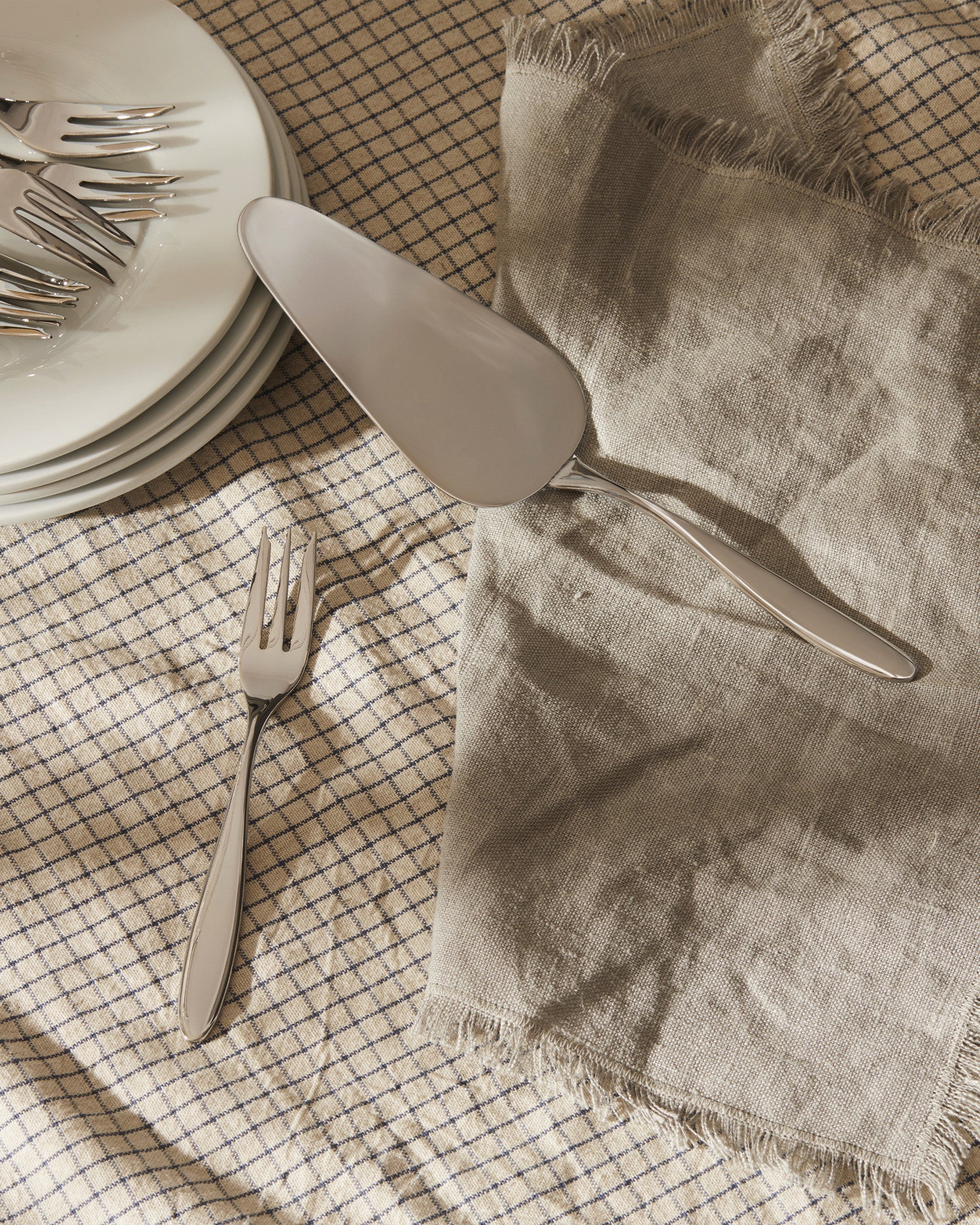 Sleek silver cake server by Alessi and a matching fork, on a checkered tablecloth with a linen napkin and stacked white plates.