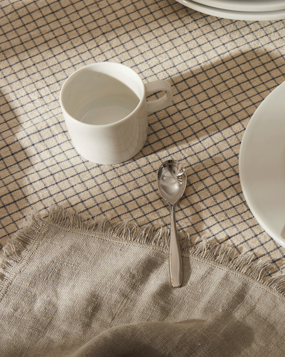White ceramic mug by Alessi on a plaid tablecloth with a silver spoon and plate.