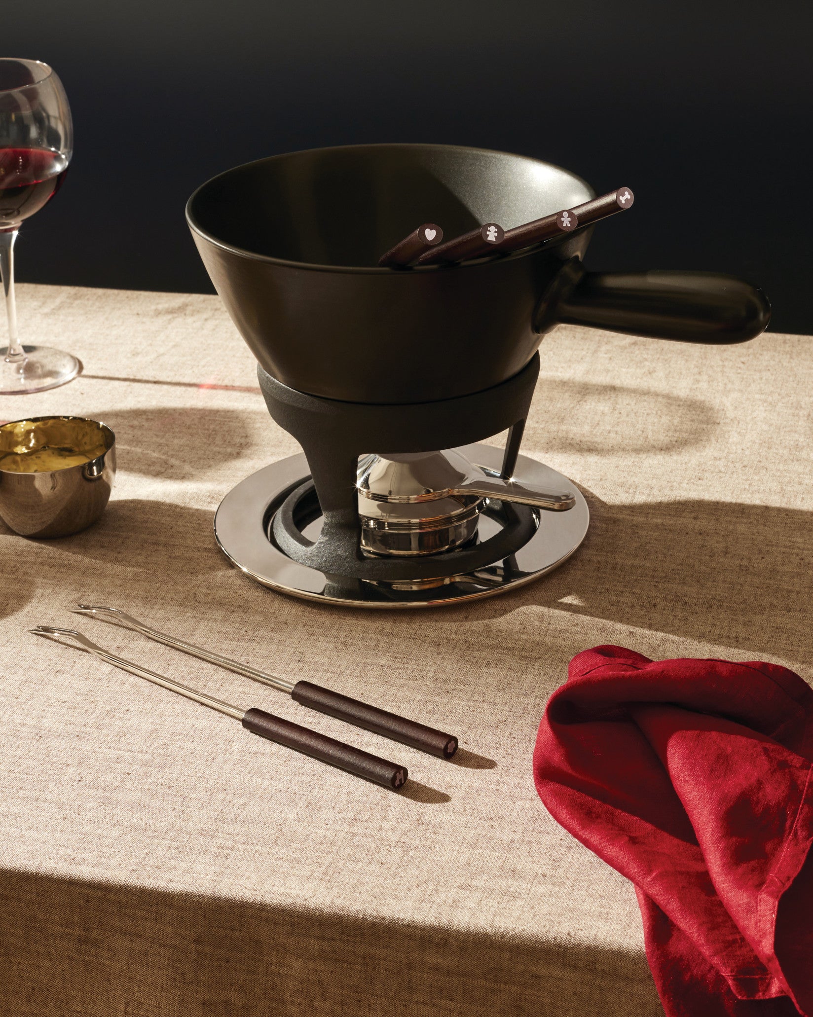 Black fondue set by Alessi with a shiny silver base, individual forks marked with symbols, and red wine.
