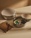 Textured silver round storage boxes by Alessi, one open containing stationery, another closed, beside a notebook with a pencil.