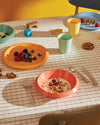 Colorful kids' dining set by Alessi, including plates, bowls, and cups, arranged on a grid tablecloth with breakfast food and toys.
