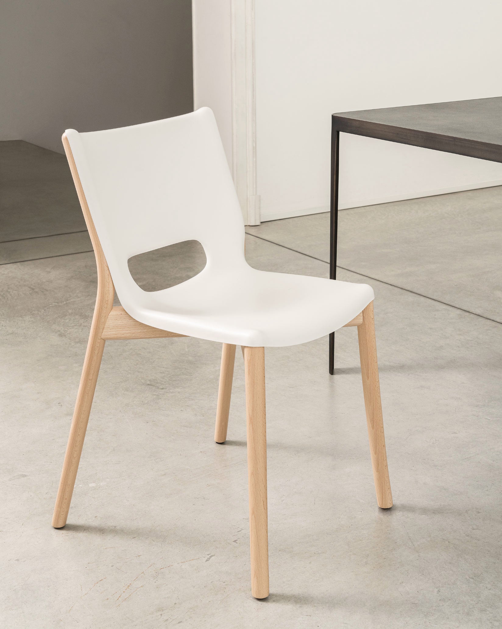 A white chair with light wood legs by Alessi, featuring a cut-out backrest, next to a dark table on a concrete floor.