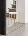 White stool with a contoured seat and four light wood legs by Alessi, on a concrete floor with granite stairs in the background.