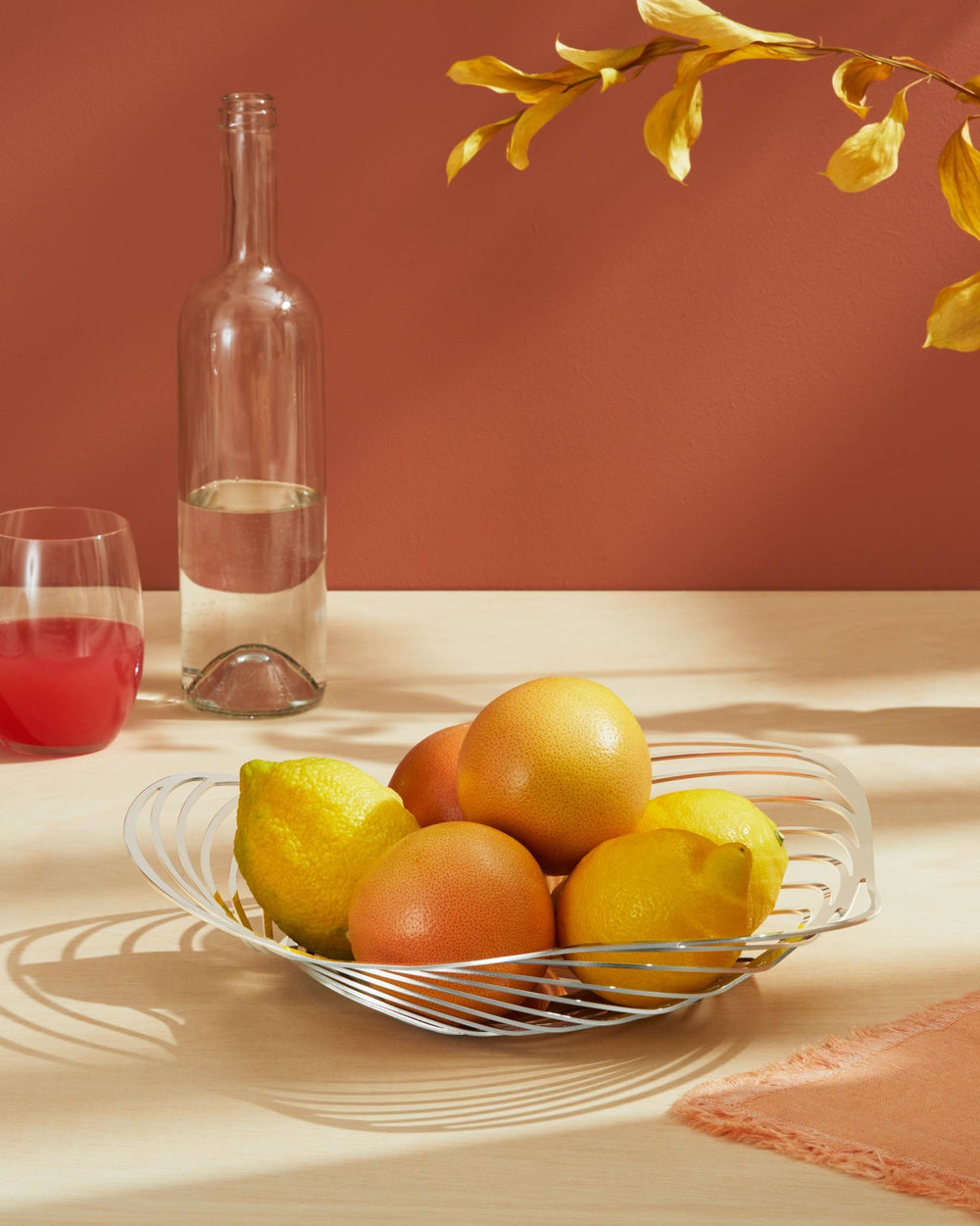 Silver-toned metallic sculptural Trinity fruit bowl by Alessi, holding lemons and grapefruits on a light wood surface.